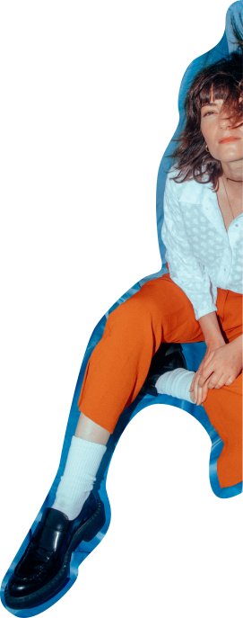 A cutout of Rowena wise sitting on a blue blanket with one leg outstretched, and her hands resting on her lap. She is wearing vibrant orange pants, a white shirt, and black loafer style shoes with white socks. She looks up and to the left, with a thoughtful expression on her face. The wind blows her hair across her face.